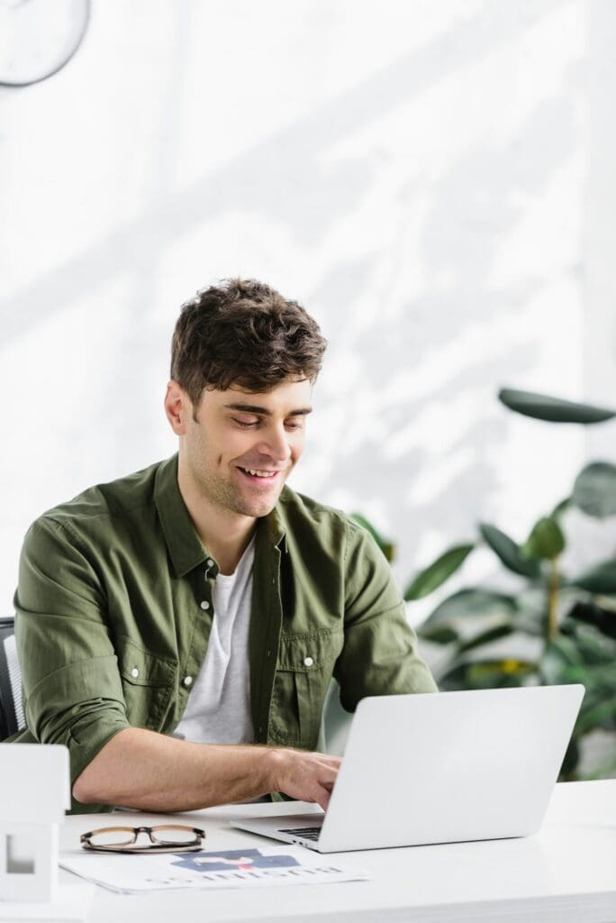 handsome architect sitting at table, typing on laptop and smiling in office