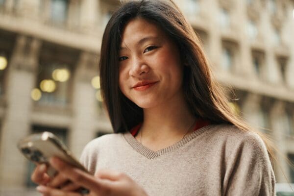 Smiling Asian woman checking her social media on smartphone looking at camera and smiling outdoors